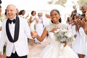 Smiling Groom in Tuxedo and Bride in Ballgown and Headpiece Walk Between Guests