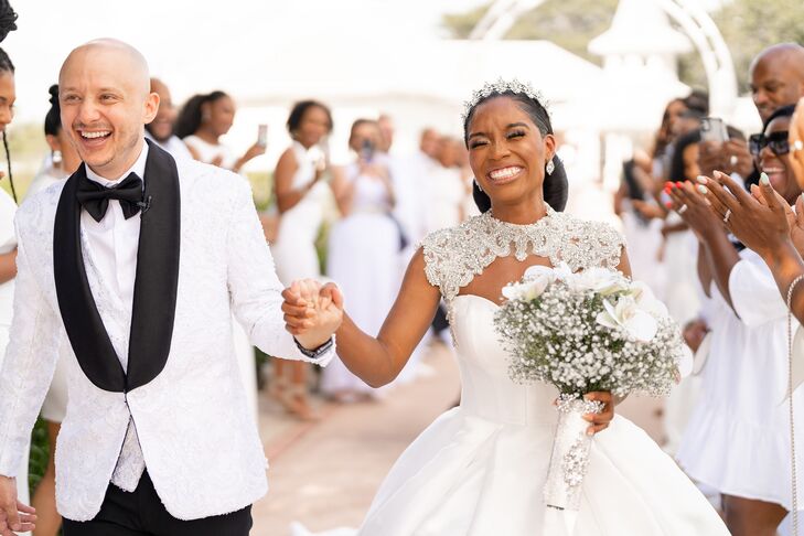 Smiling Groom in Tuxedo and Bride in Ballgown and Headpiece Walk Between Guests