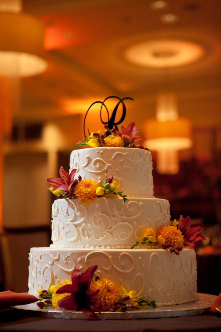 Sara and Rob's three tier wedding cake was finished with ivory frosting and elegant swirl detail and a cascade of fresh blooms in the day's color palette. A monogram letter "R" finished the confection.