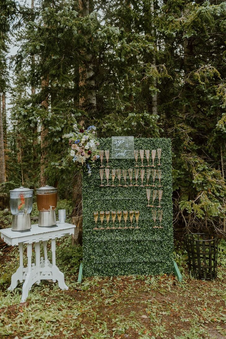 A wall of greenery displaying Champagne flutes acting as escort cards for guests to find their seats at a micro-wedding in Telluride, Colorado, with a rustic, wildflower theme amongst forest trees.