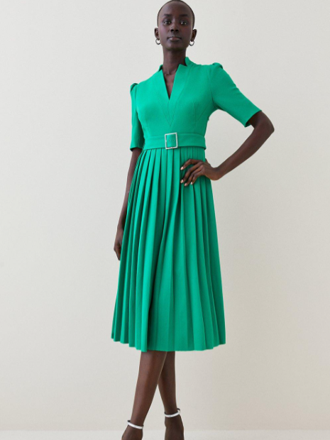A model rocks a light green wedding guest dress with a pleated skirt and belted waist.