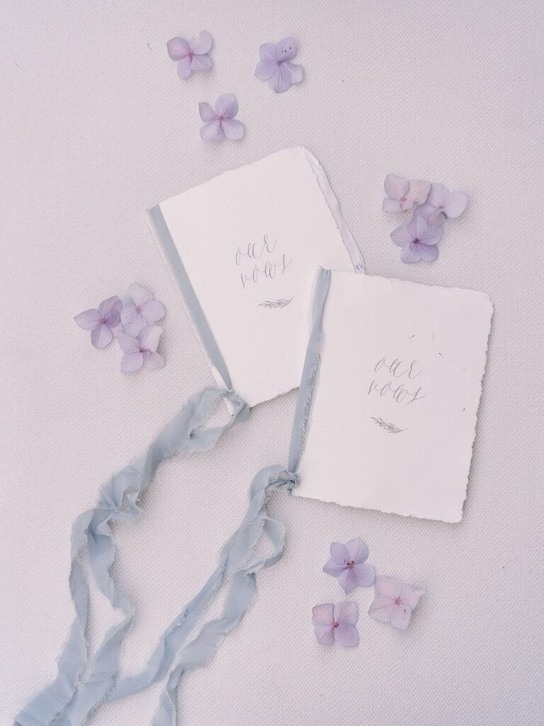 styled flat lay with handmade wedding vow books decorated with light blue silk ribbons, calligraphy and purple hydrangea petals