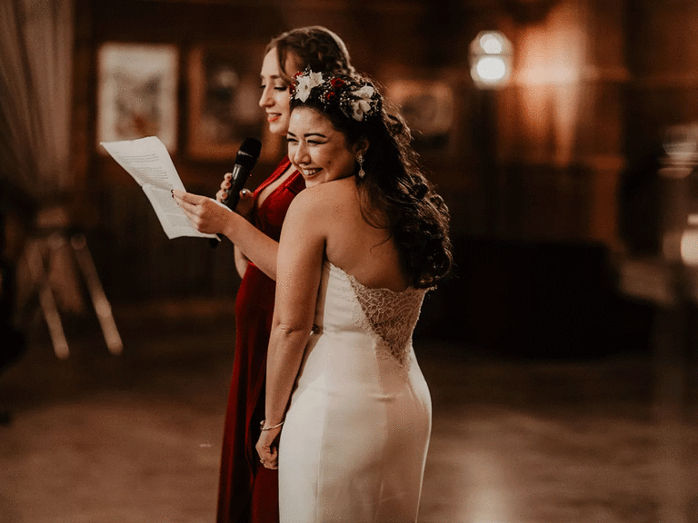 Bride stands next to maid of honor during wedding speech.