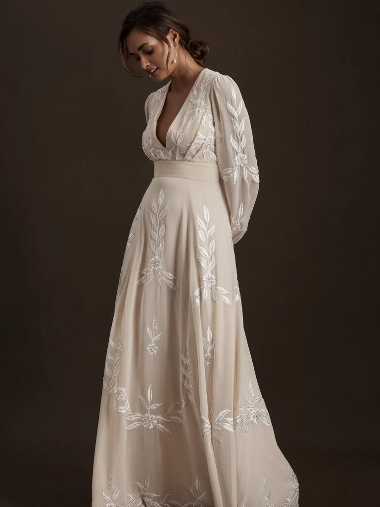 Model wears a long sleeved cream gown with floral embroidery.  