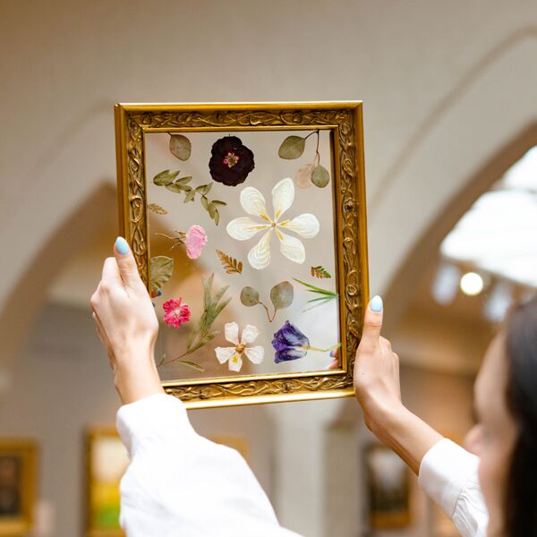 A person holds up a gilded frame of pressed flowers arranged between panes of glass.