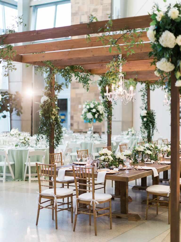 indoor wedding reception with tables under wooden pergola decorated with greenery vines and crystal chandeliers