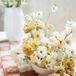 A tile-themed escort card display with guest names on terracotta tiles and a low arrangement of pastel flowers in yellow, pink, peach and white blooms.