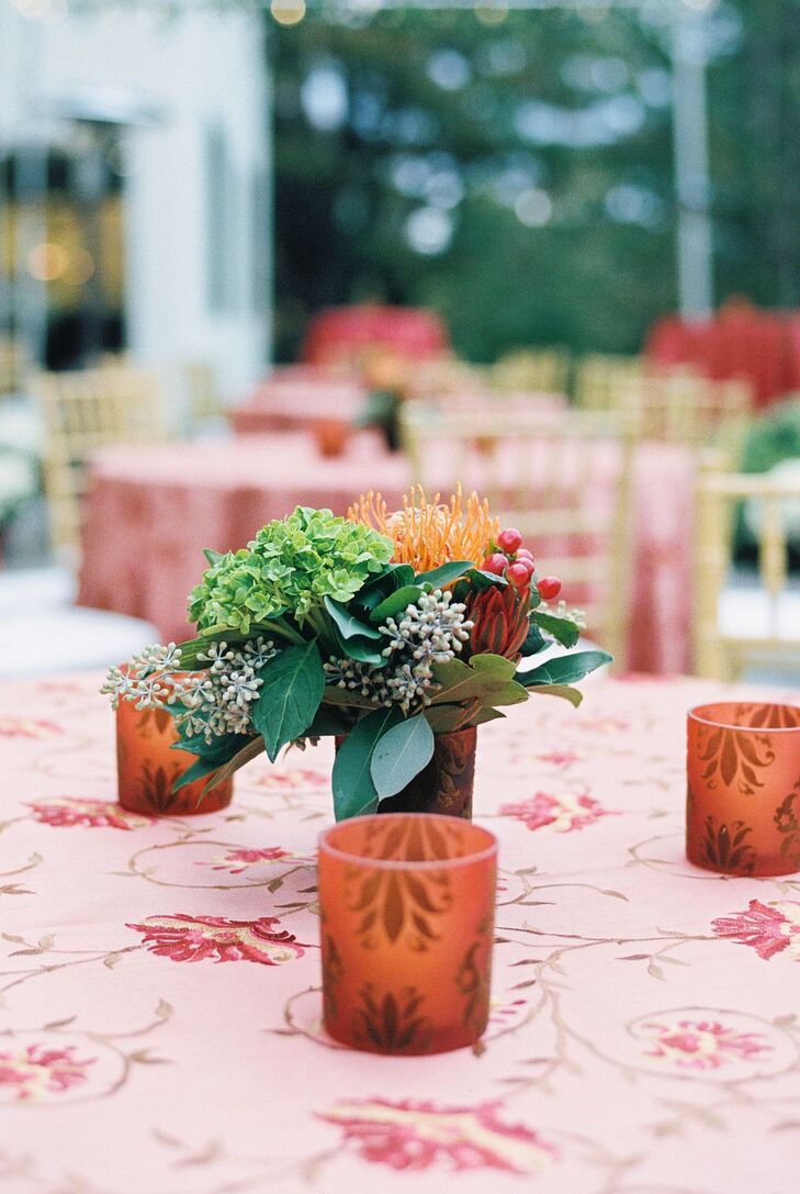 A dinner table set-up at a rehearsal welcome dinner in the Berkshires featuring a bold red linen with a floral design, low votive candles in burnt orange with a natural pattern and a low centerpiece featuring autumn flowers in orange and red accented with different shades of greenery.