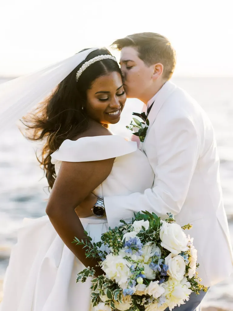 Couple in White, Suit and Wedding Dress, Kissing by the Ocean With Flowers
