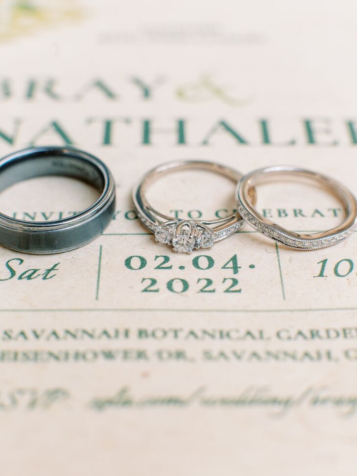 A bride's three-stone engagement ring with smaller diamond detailing, her matching wedding band, and the groom's black wedding band displayed on their invitations.