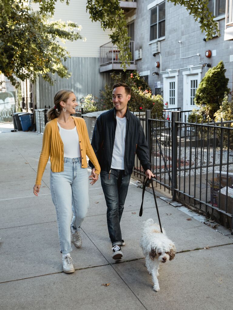 To-be-weds looking at each other while walking their dog. 