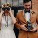 A bride in a lush flower crown and a high-neck, lace wedding dress holds binoculars while a groom in a burnt orange suit, a blue velvet bow tie and a bold boutonniere holds a vintage camera as a tribute to Wes Anderson movies at their bohemian wedding.