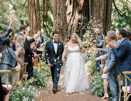 A happy couple walk back down the aisle, beaming, with their friends and towering redwoods in the background.