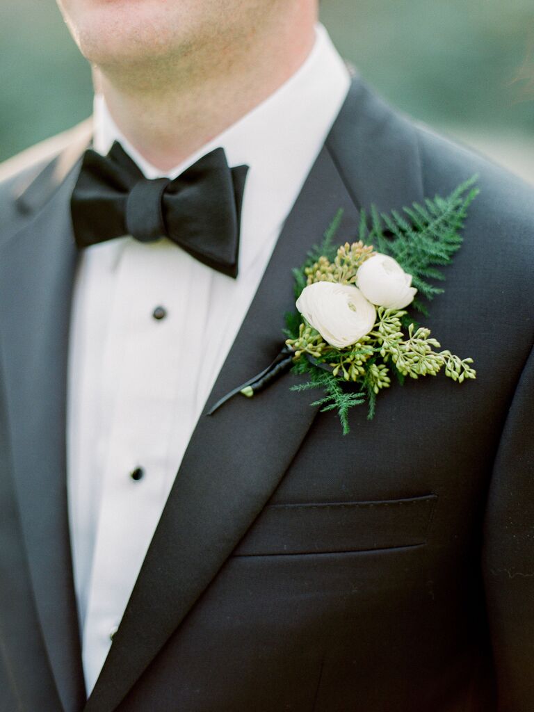 white ranunculus wedding boutonniere with greenery pinned to black tuxedo lapel