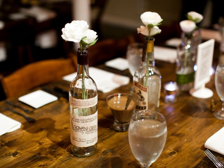 Vintage whiskey bottles hold white roses and sit on a dining table. 