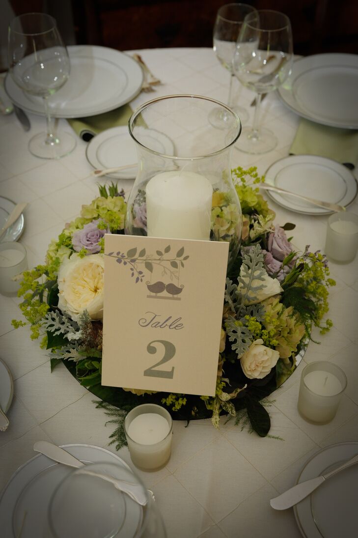 A single ivory candle was surrounded by purple and yellow roses accented with eucalyptus and greens. These pieces that embodied the natural wedding day color palette served as centerpieces for the dining tables. A table number was positioned at the front of the arrangements, which had sage green calligraphy written on ivory stationery. Smaller white candles were scattered around the centerpieces, creating a warm, welcoming glow inside the reception space.