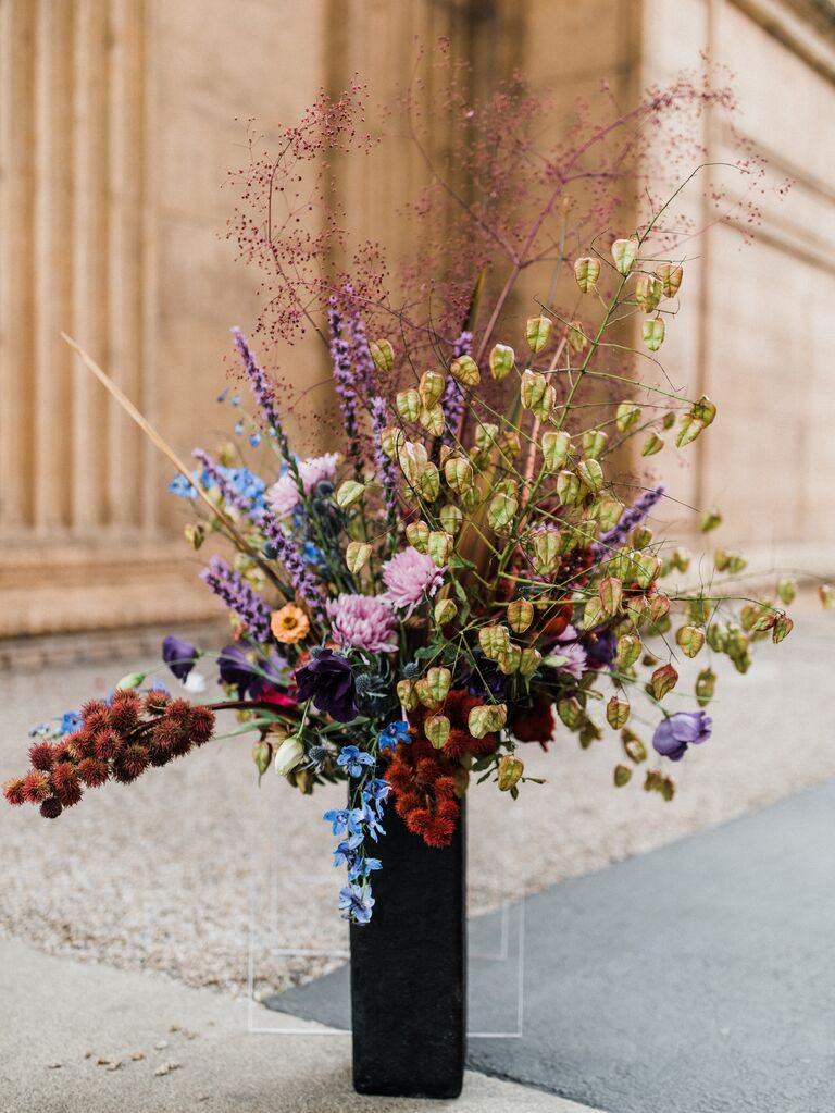 2023 wedding color trend example tall flower arrangement with dark purple, red, blue and green flowers in black rectangular vase