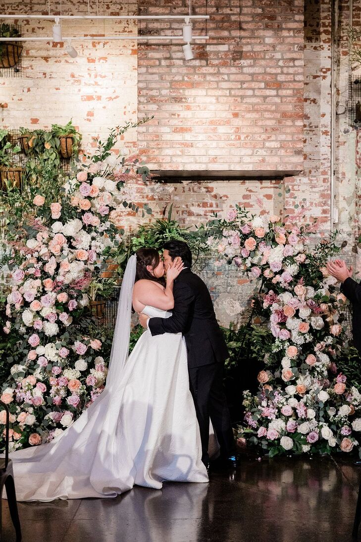 A bride in an A-line gown and a long veil and a groom in a classic black tuxedo share their first kiss as a married couple in front of an asymmetrical ceremony arch with blush, peach and white flowers in an industrial winery space.