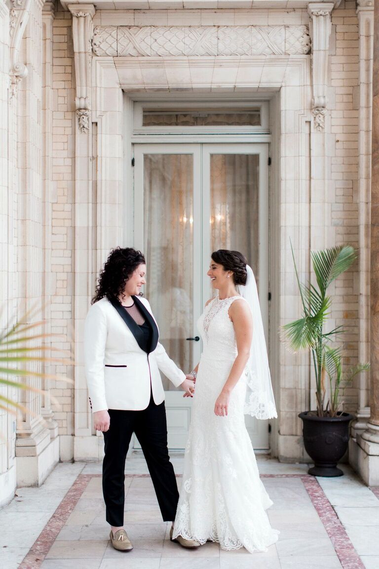 bride wearing lace wedding dress and matching fingertip length veil meets partner bride wearing black tuxedo pants and white jacket