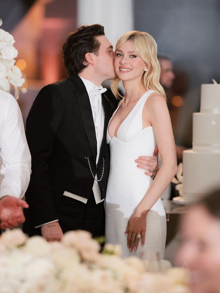 Nicola Peltz wears a chic white gown while her husband kisses her cheek. 