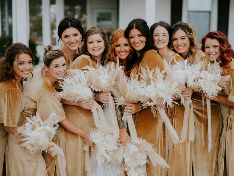 A bride poses for a photo with her wedding party, each of whom where autumnal-gold dresses and carry pampas grass bouquets.