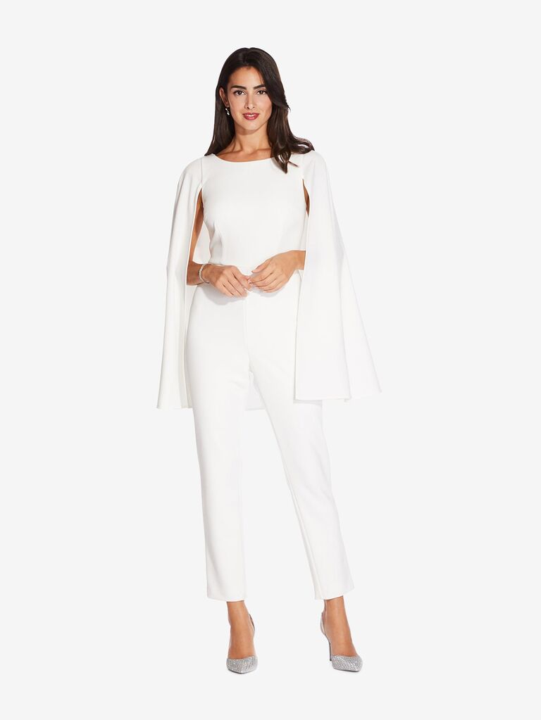 Model wears a white jumpsuit with an attached cape. 