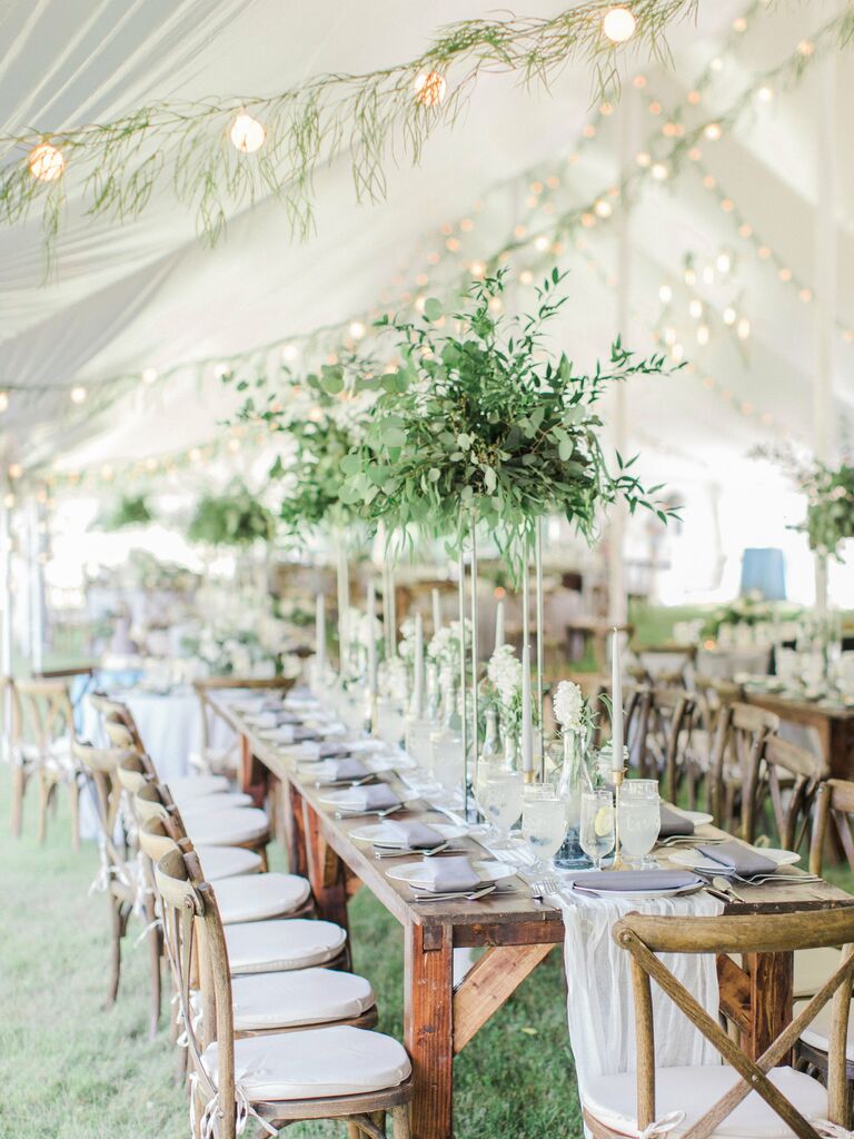 tented wedding reception with tall greenery centerpieces in the center of long wooden table