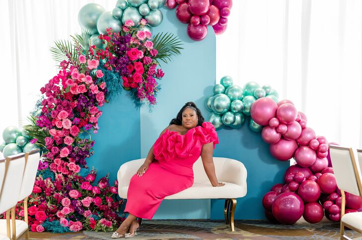 A bride-to-be in a hot pink, one-shoulder dress lounging on a white sofa at her bright and whimsical bridal shower with a custom, pale blue backdrop that featured pink, blue and purple balloons as well as bold, cascading flowers.