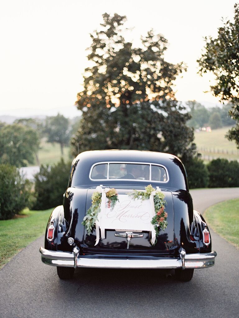 view of newlywed couple kissing through the back window of black vintage car decorated with "just married" sign on the trunk