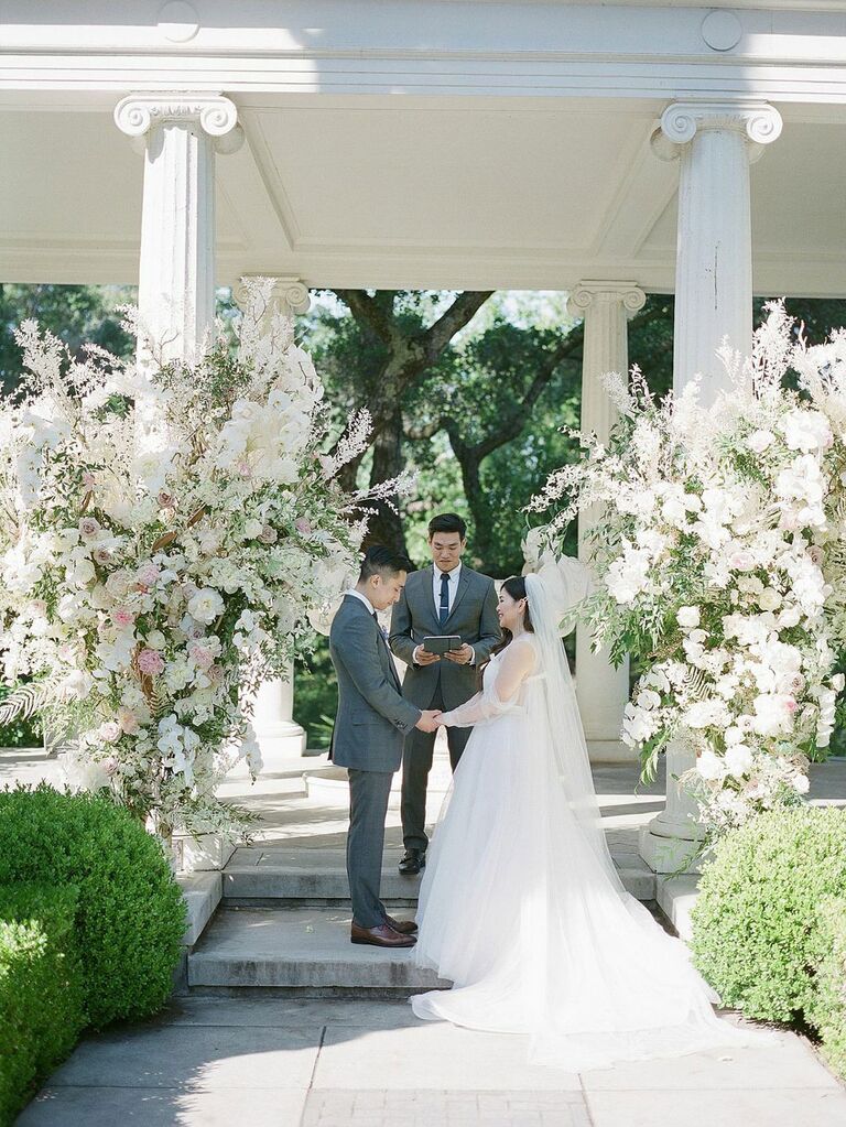 Bride and groom reciting vows in front of lavish white floral installations at summer wedding