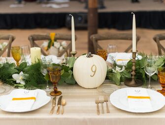 fall wedding tablescape with greenery table runner, white taper candles and white pumpkin table number with 9 in gold lettering