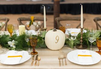 fall wedding tablescape with greenery table runner, white taper candles and white pumpkin table number with 9 in gold lettering