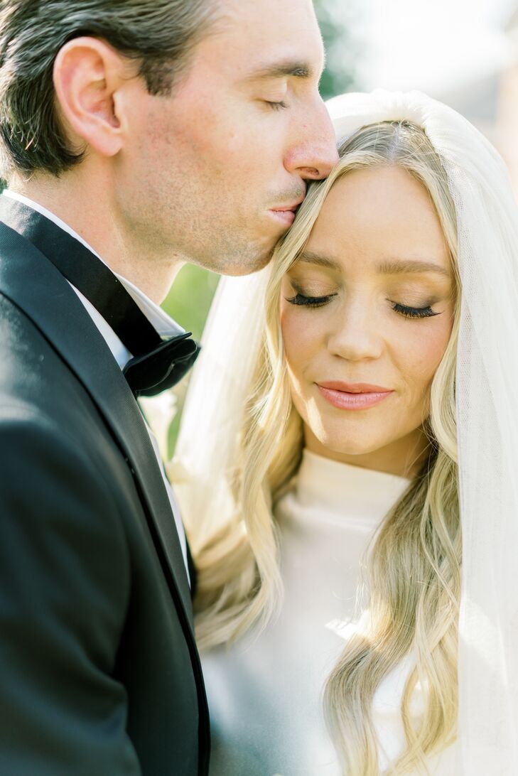 Groom in Classic Black Tuxedo Kissing Bride in High-Neck Gown, High Veil, Simple Makeup