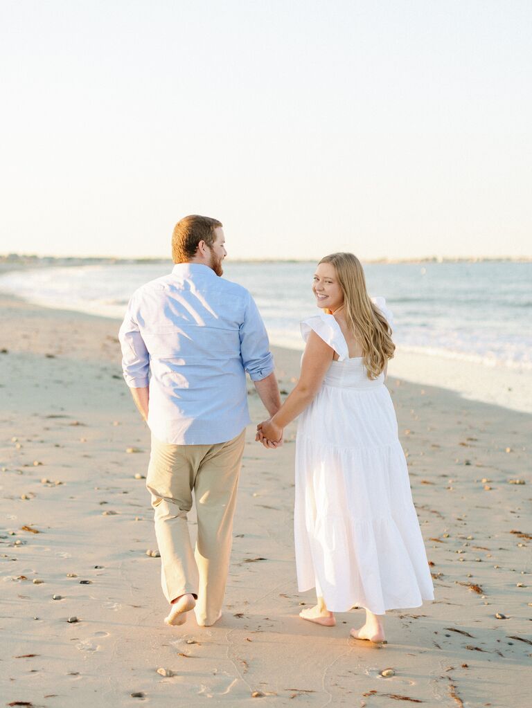 Couple walks down a beach while the bride looks back at the photographer. 