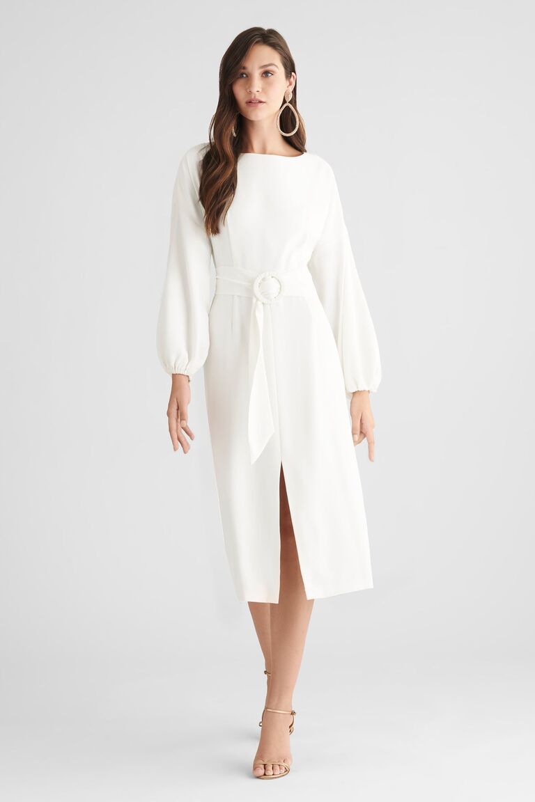 Model wears a chic white dress with long sleeves and a belted waist. 