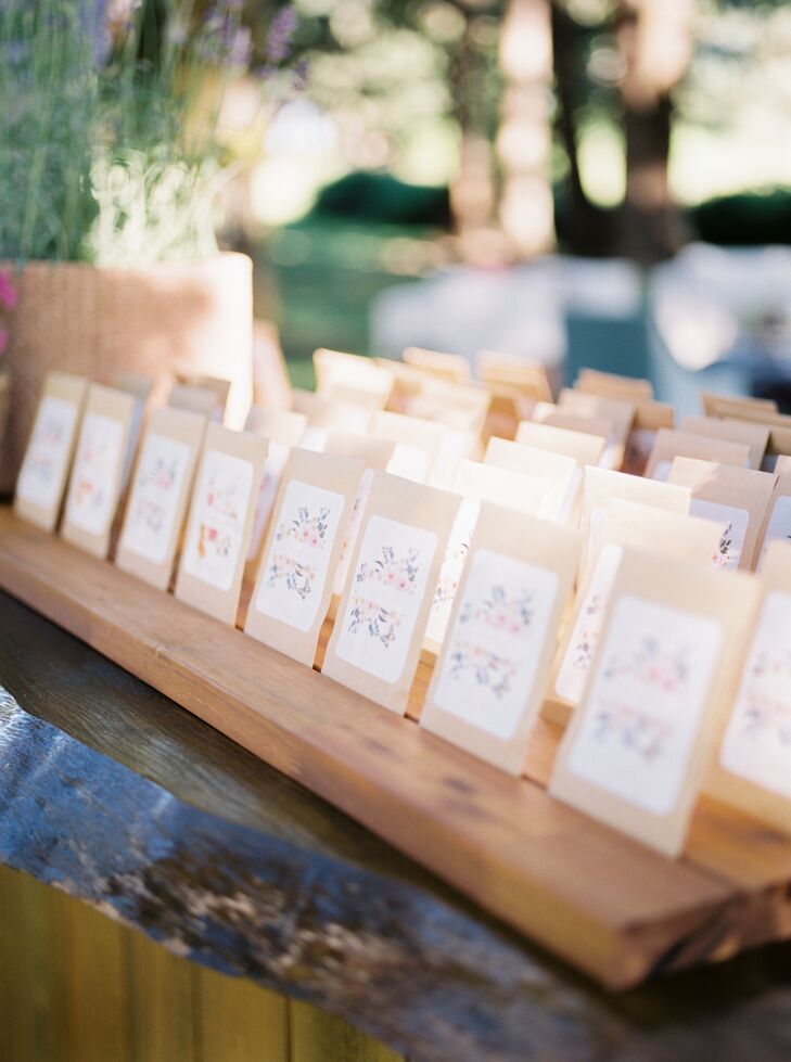 Packets of Oregon wildflower seeds as colorful, whimsical escort cards with painted flowers resting on the wooden bar at an outdoor, tented reception in the summer.