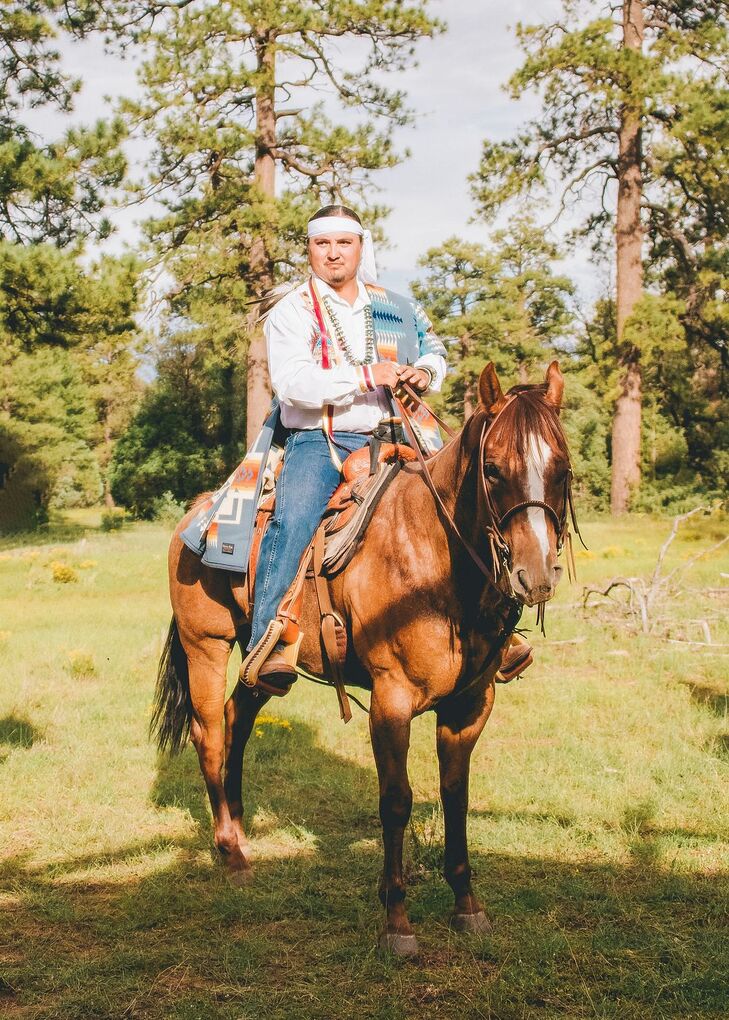 A Native American Navajo groom in headband, handmade shirt and jeans rides into his traditional wedding ceremony on horseback.
