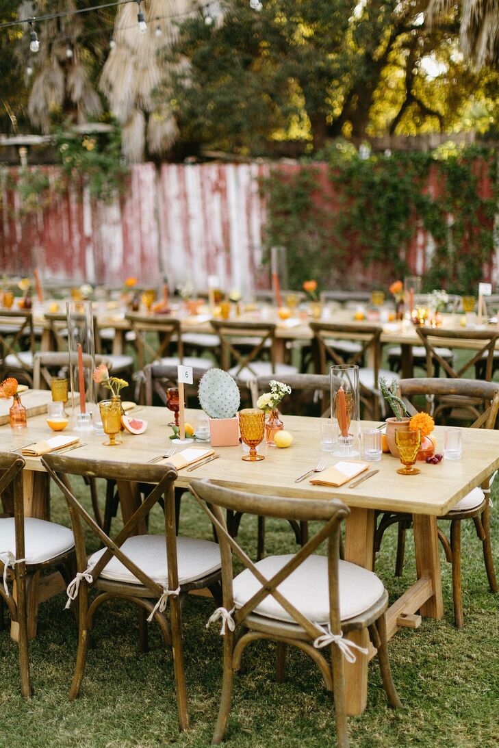 Long wooden dining tables at a casual motel backyard wedding in Ojai, California, featuring burnt orange glassware and candles, potted cacti, wildflowers and vases accented with fruits and simple linen napkins.