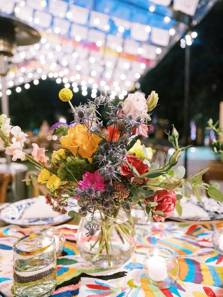 A reception dining table with a low centerpiece arrangement with light-colored blooms, low candles and a colorful patterned table linen with a bold Mexican-inspired design, all under string lights at a wedding reception in San Miguel de Allende, Mexico.