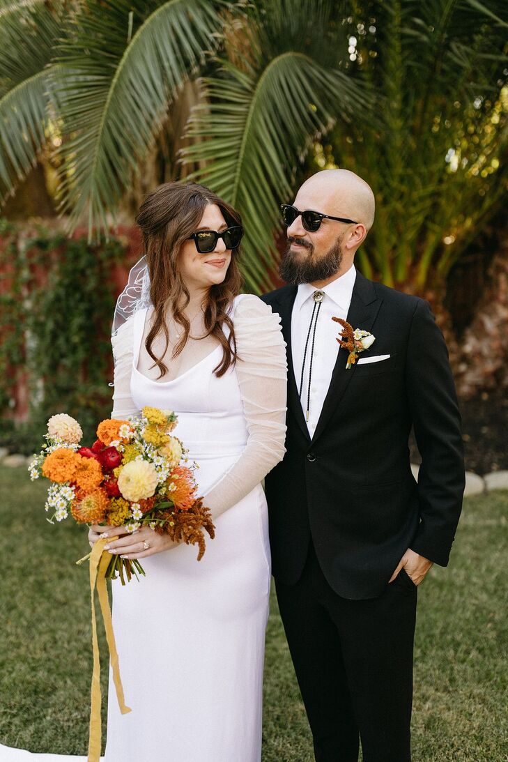 A bride in a modern Sarah Seven wedding dress with illusion sleeves and minimalistic jewelry, sunglasses, short veil and carrying a bouquet of red, yellow and burnt orange flowers standing next to her groom in a black suit and bolo tie: both wearing sunglasses at their casual, desert-inspired motel wedding.