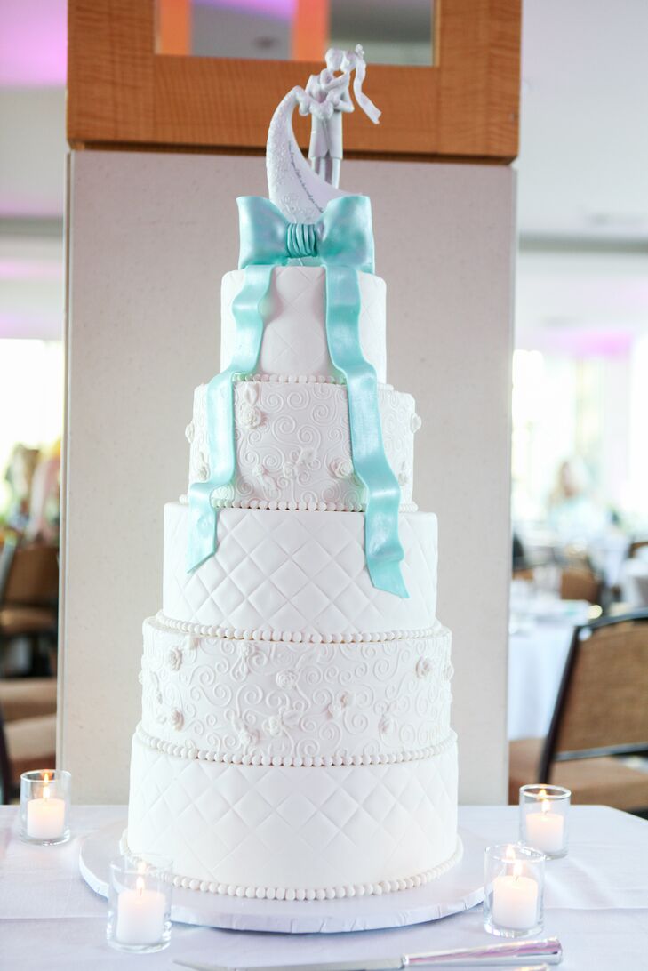 A Tiffany blue fondant bow with cascading ribbons added a graceful pop of color to this tall, elegant five-tier wedding cake. The modern white tiers alternated between quilted textures and detailed spirals with delicate flowers.