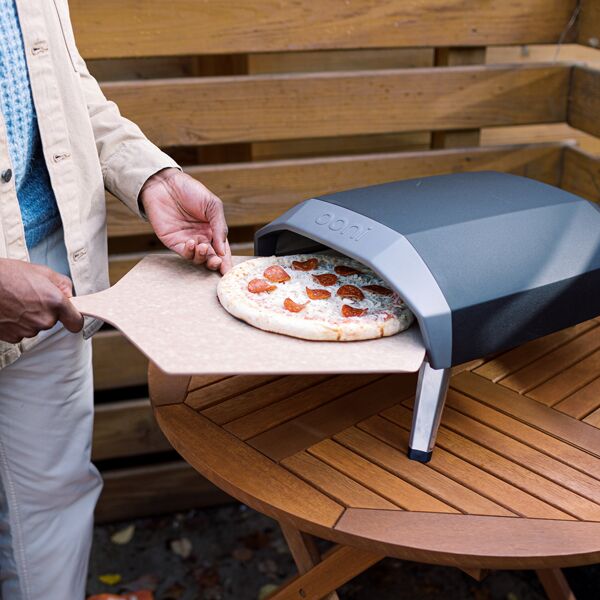 A person slides a small pizza into a fancy-looking pizza oven in a small backyard space.