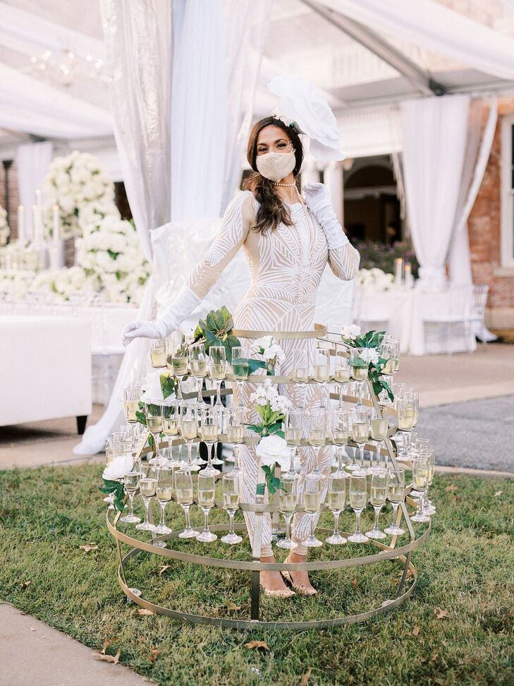 A woman in a glittering suit and gloves walks around a luxury, tented wedding reception with a tiered, structured skirt featuring glasses of Champagne for the guests.