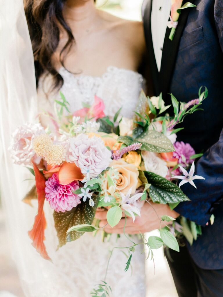 Couple holds a boquet of pastel-colored flowers, including dahlias, roses, and greenery. 