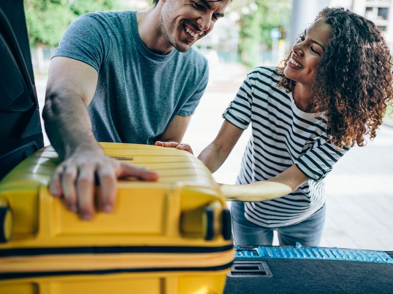 Newlyweds placing yellow hard-shell luggage in trunk 