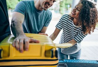 Newlyweds placing yellow hard-shell luggage in trunk 