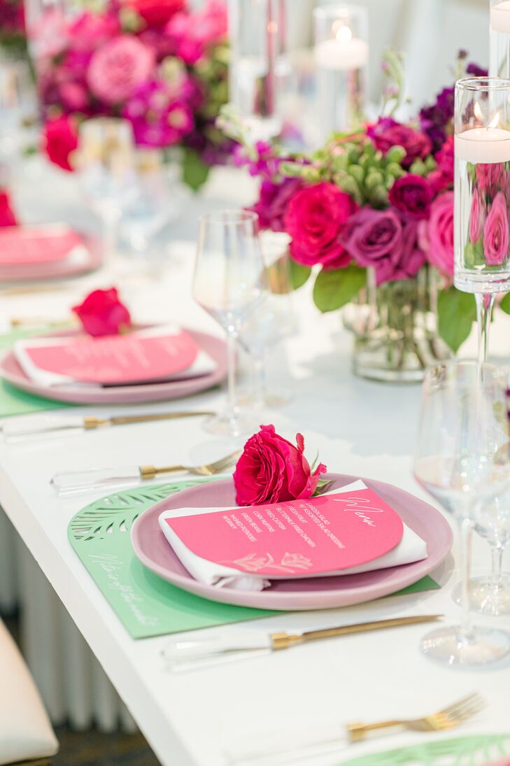 A white table with place settings featuring turquoise green placemats, light pink chargers, gold flatware, a pink rose and a hot pink dinner menu on a table with low, colorful centerpieces and glass candles.