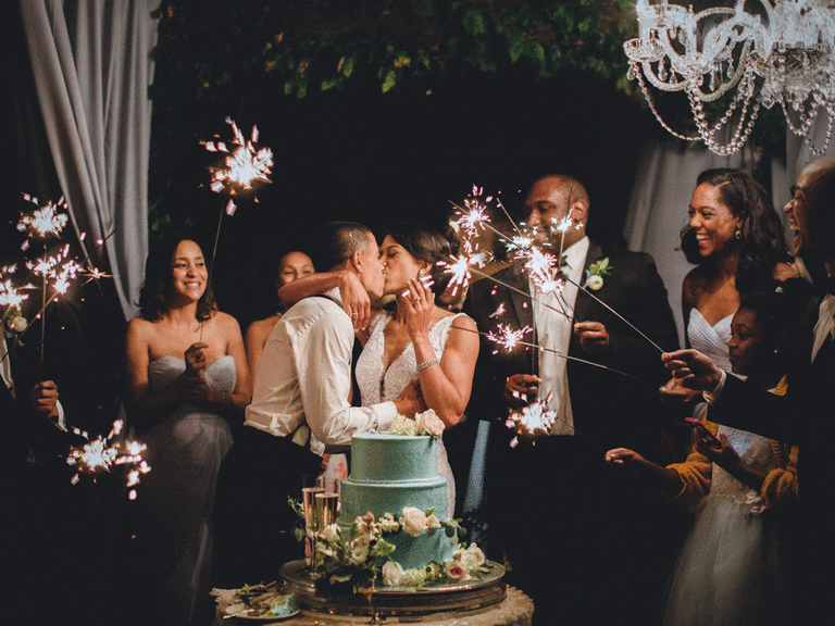 Newlyweds kissing next to wedding cake while guests hold sparklers.