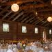 Inside the barn reception, round tables were dressed in white tablecloths with burlap runners  and decorated with wildflower centerpieces. String lights and paper lanterns hung from the ceiling of the barn venue.