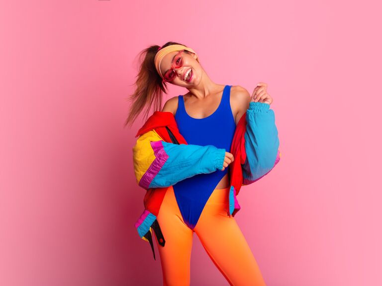 Woman wears a colorful 80s aerobic outfit in front of a pink wall. 